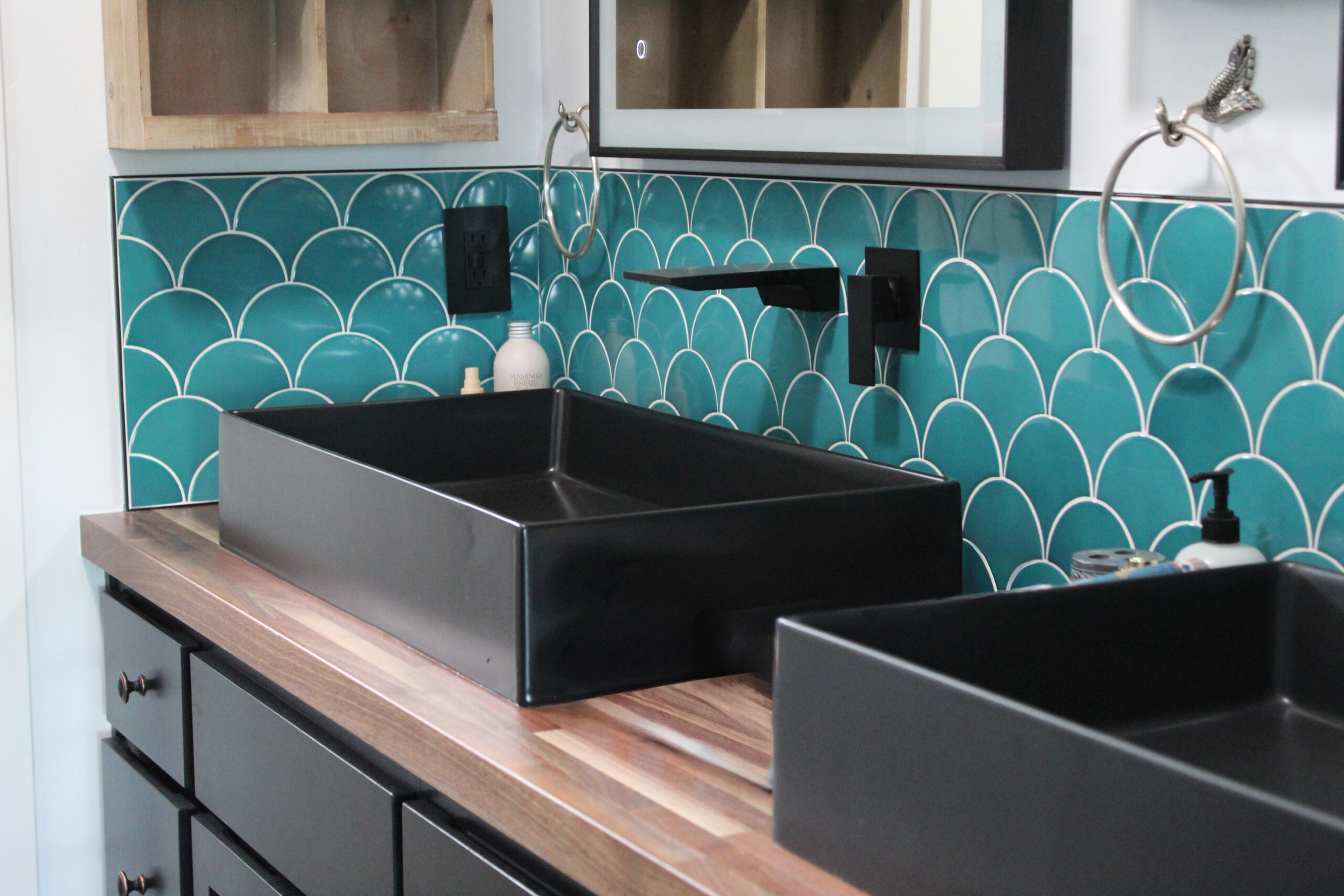 Modern bathroom vanity with dual black vessel sinks, wood countertop, matte black fixtures, and teal scalloped tile backsplash.
