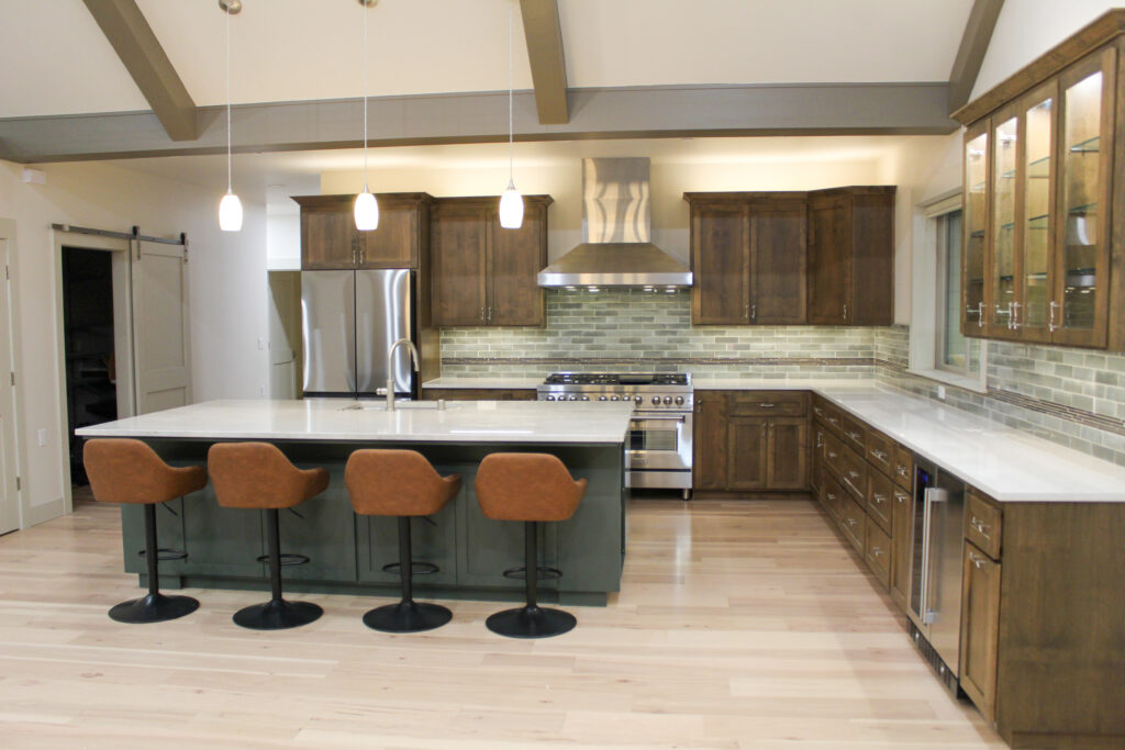 Kitchen remodel with warm wood cabinets, white countertops, green island with four leather barstools, stainless steel appliances, glass tile backsplash, and exposed ceiling beams.